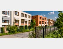 Mehrfamilienhäuser in einer modernen Wohnsiedlung mit hellen Fassaden, großen Fenstern und Markisen; Vorgärten mit Begrünung und ein Weg, der von einem Metallzaun flankiert wird, führen entlang der Gebäude. Im Hintergrund zeigt sich ein klarer Himmel mit vereinzelten Wolken.