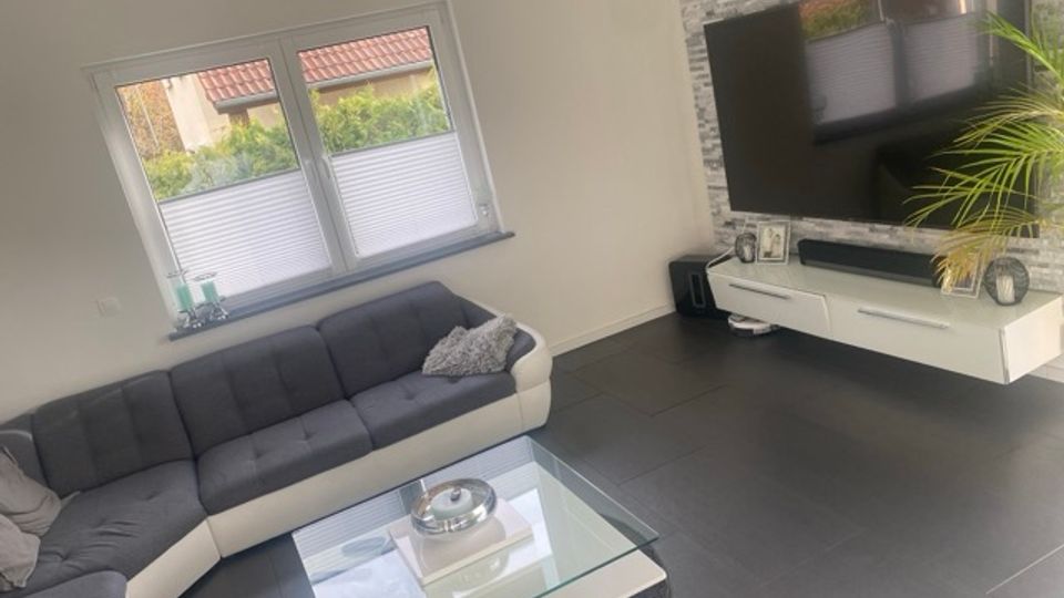 Modernes Wohnzimmer mit großem Ecksofa vor einem Doppelfenster mit Plissees sowie einem Glas-Couchtisch auf dunklen Fliesen. Gegenüber befindet sich eine TV-Wand mit grauer Steinoptik, wandmontiertem Fernseher und weißem Lowboard, daneben eine Zimmerpflanze.