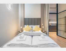 Modernes Schlafzimmer mit Doppelbett, hohem grauem gepolstertem Kopfteil, gelben Kissen und Nachttischen mit Leselampen. Rechts führt eine Glas-Schiebetür mit Vorhang in eine kleine Küche mit dunklen Schränken und Holzfußboden.