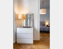 Weiße Kommode mit drei Schubladen, darauf ein rechteckiger Spiegel mit verziertem Rahmen, daneben eine Stehlampe mit hellem Lampenschirm. Im Hintergrund ist ein Raum mit einem Bett und einer weiteren Lampe zu sehen, der Boden besteht aus hellem Fischgrätparkett.