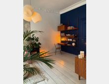 Wohnzimmer mit hellen Holzdielen, einer dunkelblauen Wand und einer Wandregal-Kombination aus Holz. Im Raum befinden sich eine große Zimmerpflanze, ein kleiner Tisch aus Holz, eine Pendelleuchte mit Lampenschirmen aus Papier sowie ein schlichter Stuhl an einem kleinen Schreibtisch.