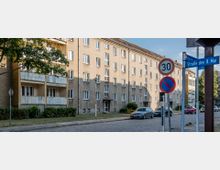 Mehrstöckiges Wohngebäude mit verputzter Fassade und kleinen Balkonen, das an einer mit Kopfsteinpflaster ausgelegten Straße liegt. Im Vordergrund sind Verkehrsschilder zu sehen, darunter ein Tempo-30-Schild und ein Halteverbotsschild, sowie parkende Autos entlang der Straße.