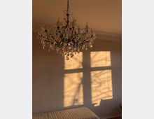 Ein Kristalllüster hängt von der Decke in einem Raum mit beigefarbenen Wänden, auf die durch ein Fenster die Schatten von Bäumen und das Fensterkreuz fallen. Darunter ist ein gepolstertes Möbelstück sichtbar.