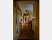 Schmaler Hausflur mit Fischgrät-Holzboden, cremefarbenen Wänden und einer Deckenleuchte. Links stehen Türen mit Milchglas, rechts ein Einbauschrank und der Beginn einer Holztreppe; am Ende führt eine offene Tür in einen weiteren Raum.