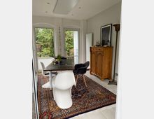 Essbereich in einem modernen Raum mit großen Fenstern und Blick auf Bäume. Der Bereich umfasst einen Holztisch mit weißen und grauen Stühlen, einen farbenfrohen Orientteppich und einen antiken Holzschrank an der Wand.