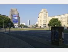 Straßenkreuzung mit breiten Fahrbahnen und Gehwegen, umgeben von mehrstöckigen Gebäuden im klassischen Architekturstil der Nachkriegszeit; im Hintergrund ein modernes Hochhaus. Ein Baugerüst mit Werbung ist an einem der vorderen Gebäude sichtbar, Bäume und Grünflächen säumen den Bereich.
