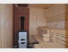 Holzverkleideter Saunaraum mit einer roten Ziegelwand, einem Saunaofen mit schwarzem Schornstein und gestapelten Steinen. Rechts befinden sich Holzbänke und ein Eimer mit Kelle, der Boden ist mit schwarz-weißen Fliesen gestaltet.