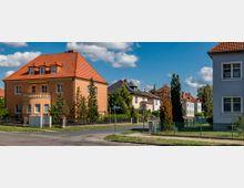 Straßenecke einer Wohnsiedlung mit mehreren zweigeschossigen Häusern, die rote Ziegeldächer und teils pastellfarbene Fassaden haben, umgeben von Vorgärten mit Bäumen und Zäunen; im Hintergrund Bäume und ein blauer Himmel mit Wolken.