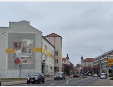 Straßenszene in einer Kleinstadt mit mehreren mehrstöckigen Gebäuden, darunter ein Gebäude mit einem großen Wandbild, das historische Bilder und Texte zeigt. Im Hintergrund sind Wohnhäuser mit roten Dächern und Zwiebeltürmen zu sehen, entlang einer befahrenen Straße mit Autos und Verkehrsschildern.
