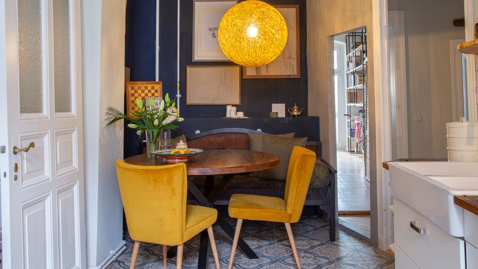 Gemütliche Essecke in einer Küche mit rundem Holztisch, zwei gelben Polsterstühlen und einer eingebauten Sitzbank vor einer dunkelblauen Akzentwand mit gerahmten Bildern. Über dem Tisch hängt eine große runde Pendelleuchte, und der Boden ist mit gemusterten Fliesen ausgelegt; rechts ist ein weißes Spülbecken und ein Durchgang zu einem Flur zu sehen.