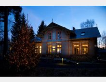 Ein zweigeschossiges Haus im Fachwerkstil mit beleuchteten Fenstern, umgeben von Bäumen und Dämmerlicht. Im Vordergrund steht ein geschmückter und beleuchteter Weihnachtsbaum.