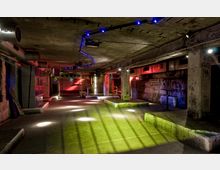 Industrieartig gestalteter, unterirdischer Raum mit Betonwänden, Pfeilern und sichtbaren Lüftungsrohren an der Decke. Farbiges Licht in Blau, Rot und Grün sowie Lichtstrahlen erzeugen eine atmosphärische Beleuchtung; die Umgebung wirkt wie ein verlassenes Lager oder ein Clubbereich.