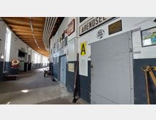 Ein langer, schmaler Flur in einem historischen Bahndepot mit Holzdecke, weißen Wänden und großen Fenstern. An den Wänden hängen diverse Schilder, Plakate und Werkzeuge, während im Raum alte Bahnutensilien, wie eine Waage und ein Wagen, zu sehen sind.