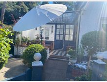 Kleiner Garten vor einem weißen Gebäude mit dunkelblauen Türen und einem schwarz-weißen Schachbrettboden im Eingangsbereich. Umgeben von Pflanzen, Topfpflanzen und einem großen weißen Sonnenschirm; im Vordergrund befinden sich dekorative Hecken und ein niedriger Zaun.