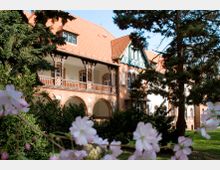 Historisches Gebäude mit Backsteinfassade und roten Dachziegeln, verziert mit Balkonen, Rundbögen und Fachwerkelementen, umgeben von grünen Bäumen und blühenden Sträuchern. Im Vordergrund sind unscharf rosa Blüten zu sehen.