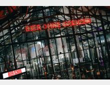 Innenansicht einer modernen Brauerei mit großen silbernen Gär- und Lagertanks, die hinter einer Glasfassade sichtbar sind. Ein Leuchtschild mit der Aufschrift „Bier ohne Grenzen“ und ein weiteres Schild mit „Beer School“ sind prominent beleuchtet.