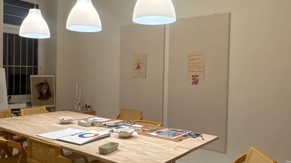 Heller Atelier- oder Seminarraum mit großem Holztisch und mehreren Holzstühlen, auf dem Tisch liegen Zeichen- und Bastelmaterialien. An der Decke hängen drei weiße Pendelleuchten und eine Schienenbeleuchtung, an der Wand sind große Pinnflächen und ein Fenster mit Gitter zu sehen.