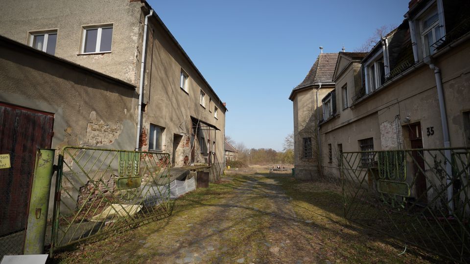 Verlassener Hofdurchgang zwischen zwei alten, stark verwitterten Wohngebäuden mit bröckelndem Putz und teils zugemauerten Fenstern. Ein schmaler, unebener Pflasterweg führt durch ein offenes Metalltor hinaus in eine grasige Freifläche unter blauem Himmel.