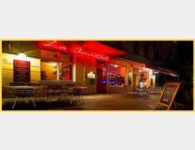 Außenansicht eines beleuchteten Cafés oder Restaurants namens "La Amistad" bei Nacht, mit roten Neonbuchstaben über dem Eingang. Vor dem Gebäude stehen Tische und Stühle auf einer gepflasterten Terrasse, daneben eine Tafel mit einem Angebot.