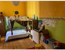 Ein farbenfrohes Kinderzimmer mit gelben und grünen Wänden, verziert mit einer Tierbordüre. Im Raum stehen ein Bett mit einem dekorativen Baldachin, ein Holzkleiderschrank, ein Regal mit Spielzeug und Büchern sowie eine kleine Kommode mit einer Orgel darauf. Der Fußboden besteht aus Holzdielen.