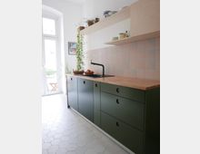 Minimalistische Küche mit moosgrünen Unterschränken, einer Holz-Arbeitsplatte und mattierten, rosafarbenen Wandfliesen im geometrischen Muster. Oberhalb befinden sich helle Holzregale mit Geschirr und Pflanzen; der Boden ist mit weißen, sechseckigen Fliesen ausgelegt, und eine Glastür führt zu einem Balkon.