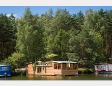 Hausboot aus Holz, das auf einem ruhigen See vor einer dicht bewaldeten Uferlandschaft liegt; mit einer Terrasse, Sonnenschirm und angrenzenden Booten. Im Hintergrund stehen hohe Bäume, hauptsächlich Kiefern und Birken.