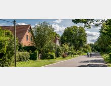 Eine ruhige Landstraße mit breitem Gehweg, gesäumt von Bäumen und Sträuchern. Links stehen Backsteinhäuser mit roten Ziegeldächern, während rechts zwei Personen auf Fahrrädern fahren.