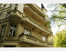 Mehrstöckiges historisches Gebäude mit verzierten Balkongeländern und reich dekorierten Fassadenelementen, umgeben von Bäumen in einer städtischen Umgebung. Ein Turm mit spitzem Dach befindet sich an der rechten Gebäudeseite.