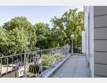 Ein Balkon mit schmiedeeisernem Geländer in einem hellen, klassischen Stil, mit Blick auf grüne, dicht belaubte Bäume. Im Hintergrund ist ein gepflasterter Weg und parkende Autos zu sehen.