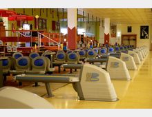 Eine Bowlingbahn mit mehreren Bahnen, ausgestattet mit Sitzreihen in grauer und blauer Farbgebung sowie automatisierten Kugelrückgabesystemen der Marke Brunswick. Im Hintergrund befinden sich eine erhöhte Sitzecke, rote Geländer, Dekorationen und Posters an den Wänden.