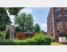 Backsteingebäude mit großen Fenstern und einer kleineren, flachen Backsteinstruktur im Vordergrund, umgeben von Bäumen und dichter Vegetation; rechts im Bild ein Industriegebäude und ein geparkter weißer Transporter.