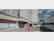 Wohnblock mit Betonfassade und niedrigen Anbauten, die teils leerstehende Fensterfronten mit roten Rahmen aufweisen. Im Vordergrund ein gepflasterter Hof, im Hintergrund weitere Hochhäuser und ein Schild mit der Aufschrift „Senioren-Freizeitstätte“.