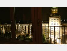 Nachtaufnahme durch ein Fenster mit Geländer im Vordergrund. Im Hintergrund beleuchtete Fassaden eines historischen Gebäudekomplexes mit klassizistischen Elementen und einem beleuchteten Turm.