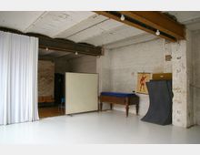 Ein einfacher Raum mit weißen Wänden und sichtbaren Backsteinen, ausgestattet mit einem weißen Vorhang, einem Holzklapptisch, stapelbaren Matten und einem Anatomieposter an der Wand. Im Hintergrund sind eine Garderobenstange, einige Stühle und ein hölzerner Boden erkennbar.