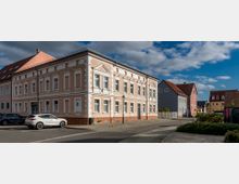 Eckgebäude im Stil der klassischen Architektur mit hellrosa Fassade, weißen Zierelementen und Sprossenfenstern; umgeben von einem gepflasterten Gehweg, parkenden Autos und angrenzenden Wohnhäusern mit roten Dächern unter einem teils bewölkten Himmel.