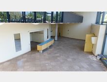 Weitläufiger Innenraum eines modernen Gebäudes mit beiger Wand- und Bodenfarbe, einer Empfangstheke aus Holz mit Glasoberfläche und großen Fensterfronten, die Tageslicht hineinlassen. Rechts sind gelbe Türen und ein geöffneter Technikschrank sichtbar; links befindet sich ein Bereich mit Kleiderhaken.