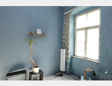 Ein schlichter Raum mit blauen Wänden, einem großen, alten Fenster mit geometrisch gemustertem Vorhang und weißem Fensterrahmen. Im Vordergrund stehen ein kleiner Beistelltisch mit einer Topfpflanze, eine schwarze Schreibtischlampe und eine weiße Stehlampe.