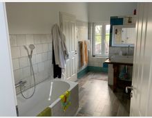 Helles Badezimmer mit gefliesten Wänden und Holzboden. Links eine Badewanne mit Handbrause, rechts ein Waschbecken auf einem Holzunterschrank, darüber ein Spiegel und ein Fenster mit Blick nach draußen.
