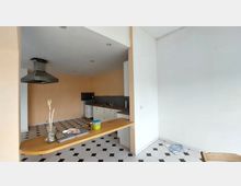 Küche in einem offenen Raum mit beige gestrichenen Wänden, schwarz-weiß gemustertem Boden und moderner Dunstabzugshaube. Die Einrichtung umfasst weiße Schränke, eine Arbeitsplatte aus Holz und eine Theke, die in den Raum ragt.