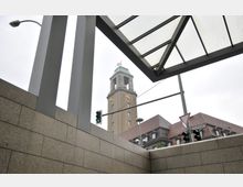 Ansicht eines Kirchturms mit einer Uhr und grünlicher Spitze im Hintergrund, aufgenommen aus einer überdachten modernen Struktur mit Metallstreben und Glasdach. Im Vordergrund sind eine gelbliche Steinwand, eine Ampel und ein Straßenschild erkennbar.