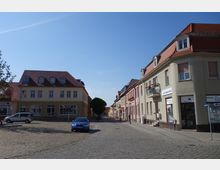 Kleine gepflasterte Straße in einer Stadt mit zweigeschossigen Häusern, die rote Schrägdächer und helle Fassaden haben. Rechts sind Geschäfte mit Werbeschildern und Blumen an Balkonen, links stehen ein Auto und ein Baum.