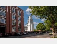 Straßenszene mit einem roten Backsteingebäude links, gekennzeichnet durch weiße Fensterrahmen, und einem modernen weißen Industriegebäude mit auffälligem Rundturm im Hintergrund. Rechts der Straße befindet sich eine Baumreihe, die Schatten spendet, und zahlreiche parkende Autos sind entlang der Straße sichtbar.
