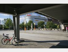 Blick von einer schattigen Unterführung auf eine städtische Kreuzung mit Bushaltestellen, Fahrradständern und einem modernen, verglasten Gebäude mit der Aufschrift „Domäne“ im Hintergrund. Im Vordergrund steht ein rotes Leihfahrrad an einem Pfeiler.