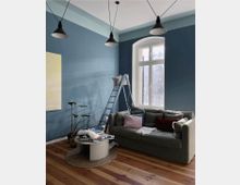 Modernes Wohnzimmer mit einer blauen Akzentwand, einer grünen Couch mit bunten Kissen, einem kleinen runden Couchtisch auf einem Teppich und einer Leiter vor einem großen Fenster. Hölzerner Boden und drei schwarze Hängelampen an der Decke.