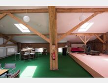 Offener Dachbodenraum mit Holzbalken, grünem Teppichboden und einer Kombination aus Wohn- und Arbeitsbereich. Links steht eine kleine Bar mit Hockern, während rechts eine Schlafgelegenheit und Fitnessgeräte auf einer erhöhten Plattform zu sehen sind; Tageslicht fällt durch zwei Dachfenster.