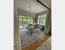 Helles Esszimmer mit großen Glasfenstern und Türen, die auf eine Terrasse mit Gartenblick führen. Ein Holztisch mit sechs Stühlen steht auf einem grauen Teppich, umgeben von hellen Wänden und einem Kronleuchter an der Decke.