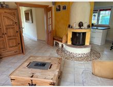 Gemütliches Wohnzimmer mit Fliesenboden, einem rustikalen Holzschrank und einem Kamin mit abgerundeter Form und dekorativen Fliesen. Der Raum führt über einen offenen Durchgang in eine angrenzende Küche mit weißen Schränken und einem Fenster.