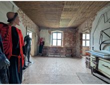 Ein rustikaler Raum mit unverputzten Ziegelwänden und Holzdecke, eingerichtet mit antiken Möbeln, wie einem alten Herd und Holzbänken. Mehrere Schaufensterpuppen, teils bekleidet, stehen im Raum verteilt, während durch zwei Fenster Tageslicht einfällt.