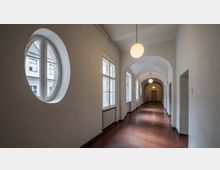 Langer, heller Flur mit gewölbter Decke, einem rötlichen Boden und runden Hängelampen. Links befinden sich ovale und rechteckige Fenster, mit Blick auf ein gegenüberliegendes Gebäude.