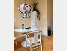 Essbereich mit modernem Design: Ein runder, weißer Tisch mit Holzstühlen steht auf einem Holzboden. Eine große, bunte Papierlampe hängt über dem Tisch, auf dem eine Glasvase und eine Schale mit Obst stehen. Im Hintergrund sind eine hohe weiße Schranktür, eine Zimmerpflanze und eine große blaue Vase sichtbar.